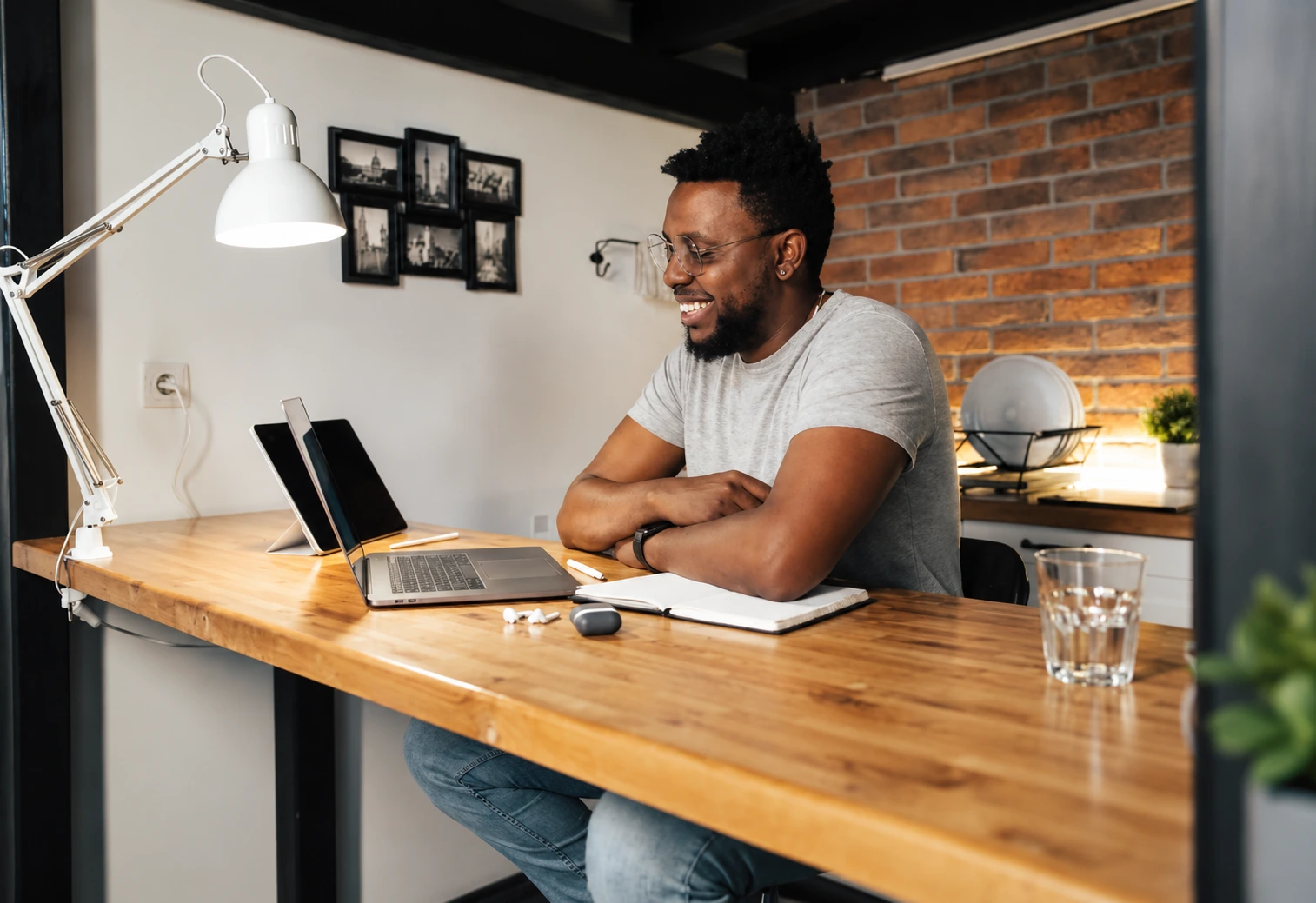 A man smiling at his laptop at a wooden desk in a brick-walled room
